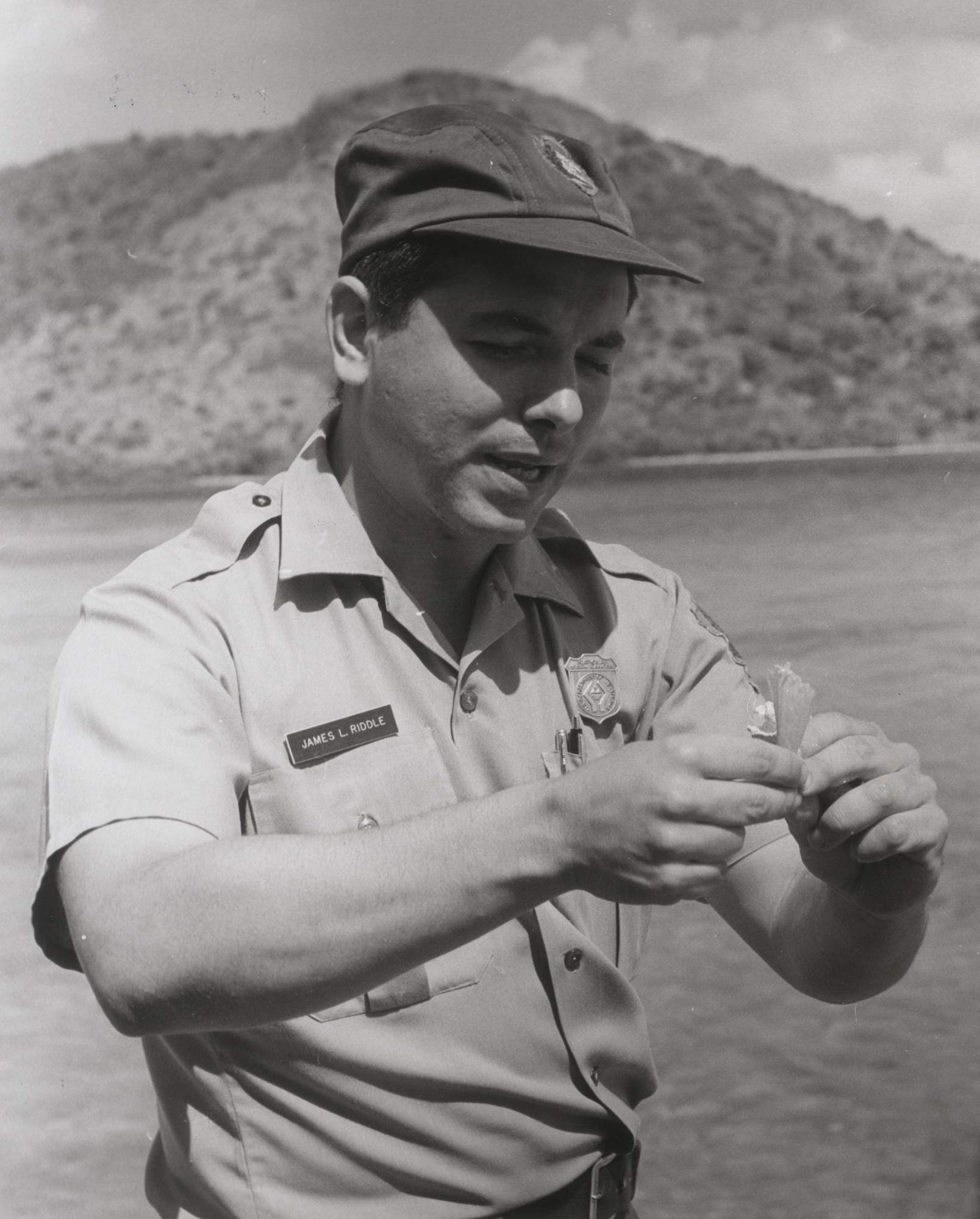 James Riddle in NPS uniform with ball cap wears a shield-shaped badge featuring stylized hands.